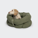 CLOUD7_ Ein kleiner cremefarbener Hund sitzt in einem runden, grünen Hundebett LTTL Snoozy Linen-Mix Moss und blickt nach links vor einem schlichten hellgrauen Hintergrund.