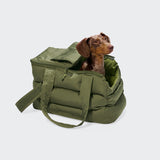Ein brauner Dackel sitzt in einer offenen, grünen, gepolsterten Hundetasche „Montreal 2.0 Light Olive“ mit Tragegriffen vor einem einfarbigen hellgrauen Hintergrund.
