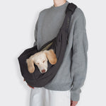 CLOUD7_ Eine Person in grauem Pullover und weißer Hose trägt einen hellen Hund in der Hundeumhängetasche LTTL Puffer Dark Grey/Khaki über der Brust. Der kuschelige Hund stützt seinen Kopf mit geschlossenen Augen auf den Taschenrand.