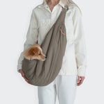 CLOUD7_ Eine Person in cremefarbenem Outfit trägt einen kleinen braun-weißen Hund in der Hundeumhängetasche LTTL Puffer Khaki/Peach über der Brust. Der Kopf des Hundes lugt seitlich vor einem schlichten weißen Hintergrund hervor.