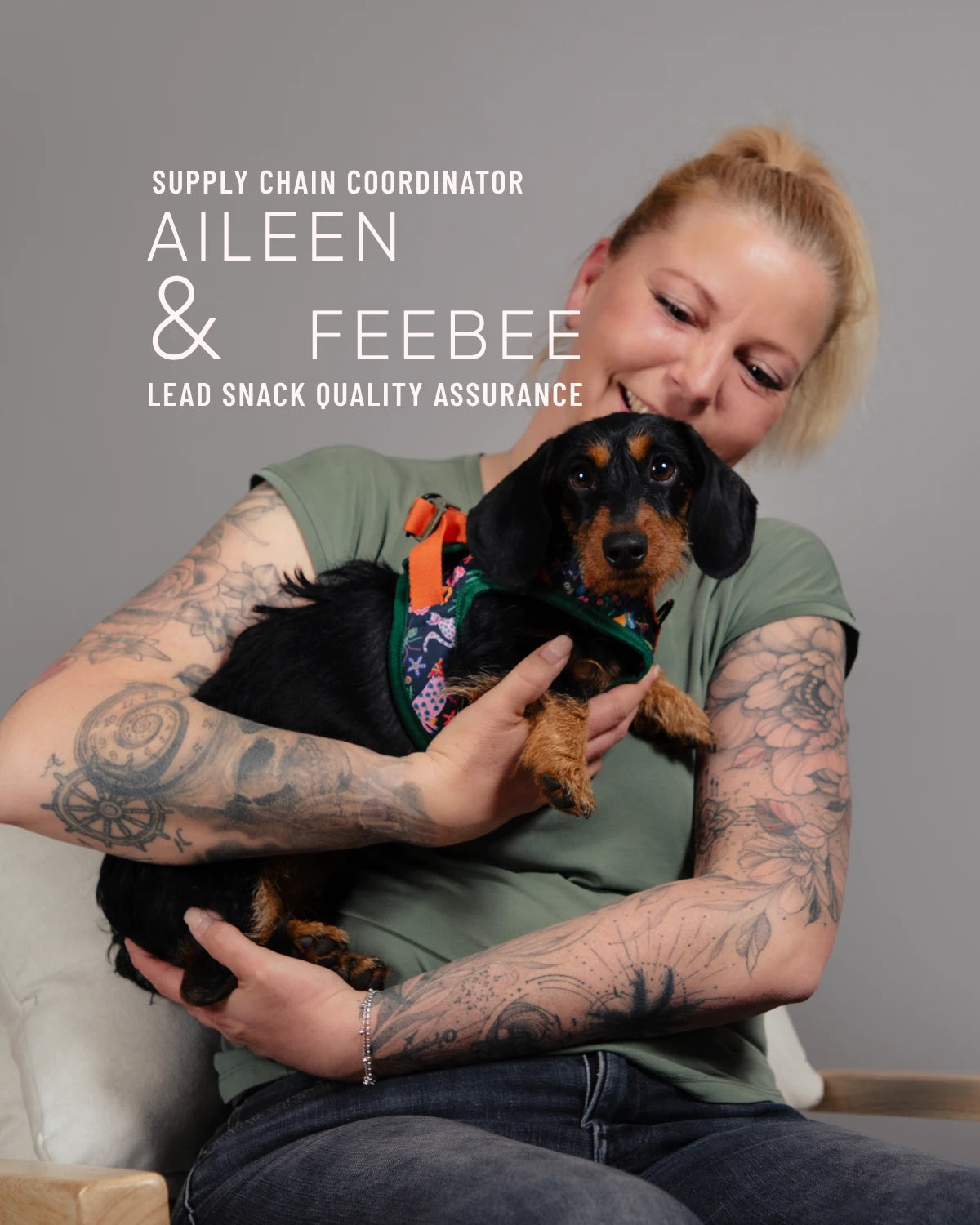 CLOUD7_ Eine Frau mit Tattoos lächelt, während sie einen schwarz-braunen Hund im Arm hält. Auf dem Bild steht: Aileen, Koordinatorin für die Lieferkette, und Feebee leiten die Qualitätssicherung für Snacks.