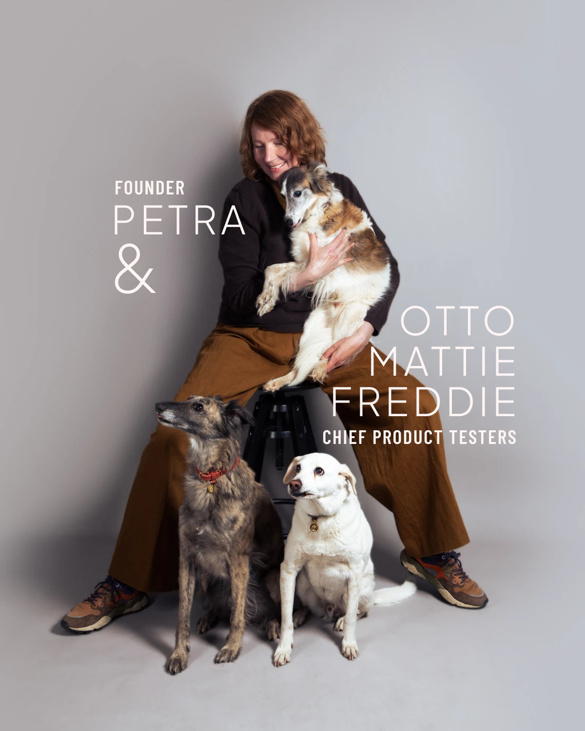 CLOUD7_ Eine Frau sitzt auf einem Hocker und umarmt einen Hund, während zwei weitere Hunde zu ihren Füßen sitzen. In der Bildunterschrift wird sie als Gründerin Petra bezeichnet, die Hunde als Otto, Mattie, Freddie und Chef-Produkttester. Schlichter grauer Hintergrund.